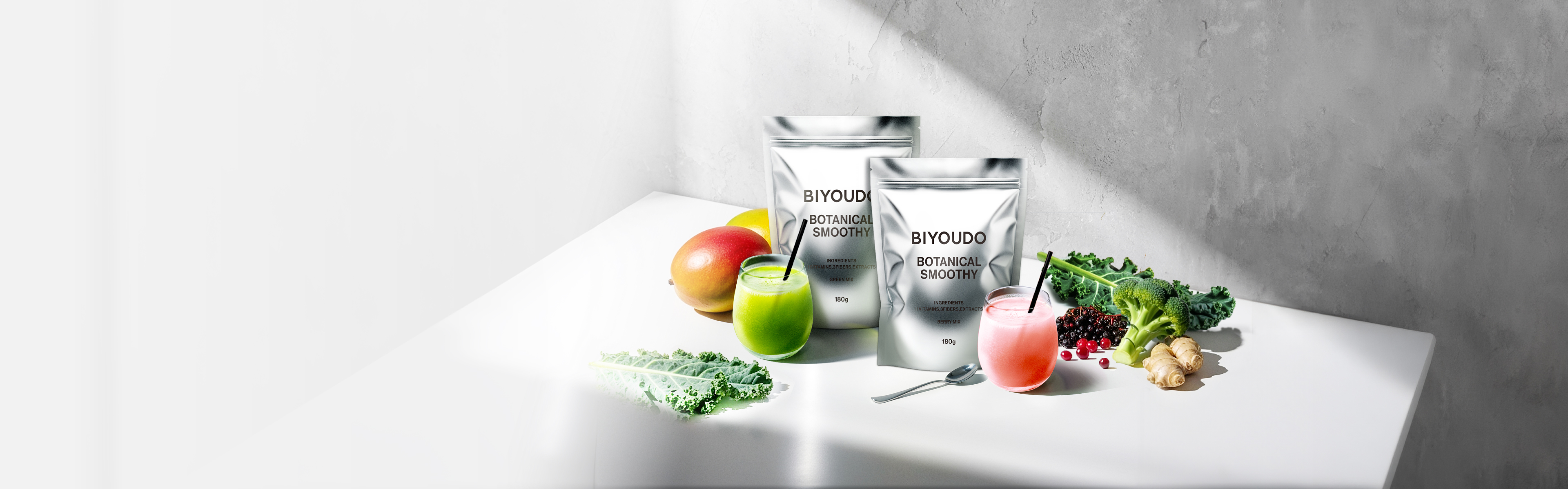 ビヨウドウ BOTANICAL SMOOTHY_白い台の上に銀色のパウチが2つ並び、緑とピンクのスムージーのグラス、マンゴーやケール、ベリーが添えられた写真